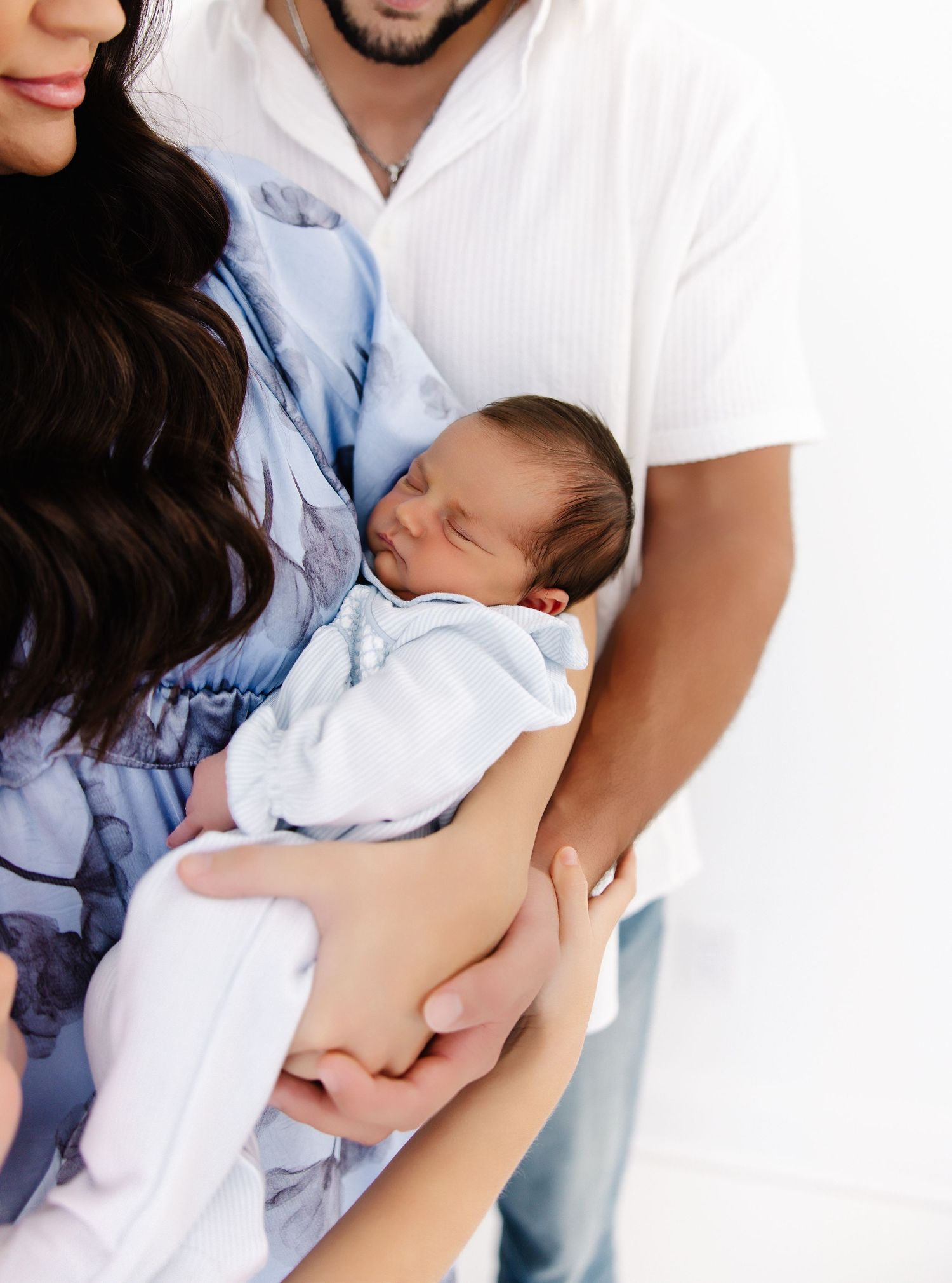 A pair of adult hands tenderly hold a sleeping newborn baby dressed in white against a white background.