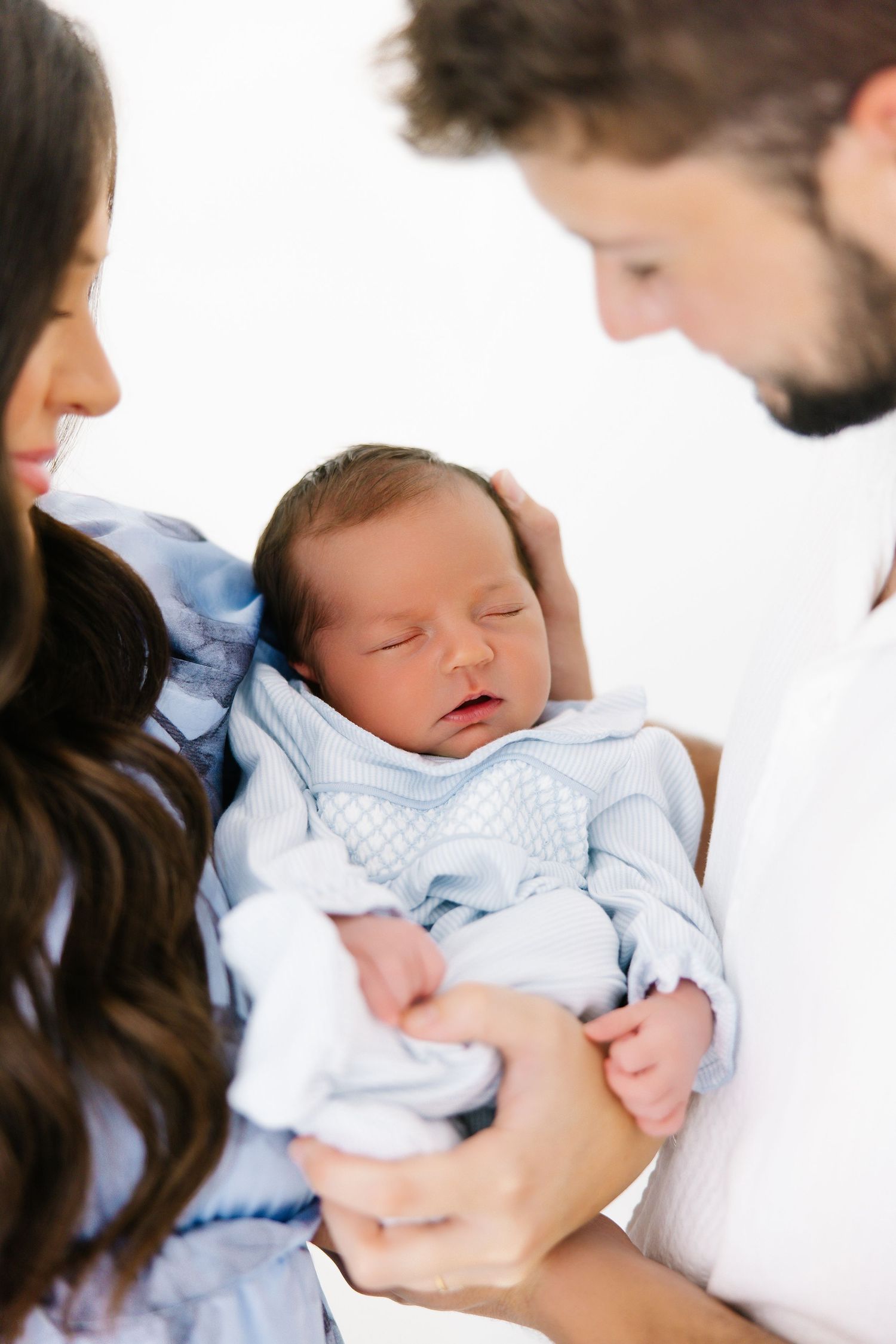 A newborn baby wearing a light blue outfit sleeps peacefully while being cradled by their parents.