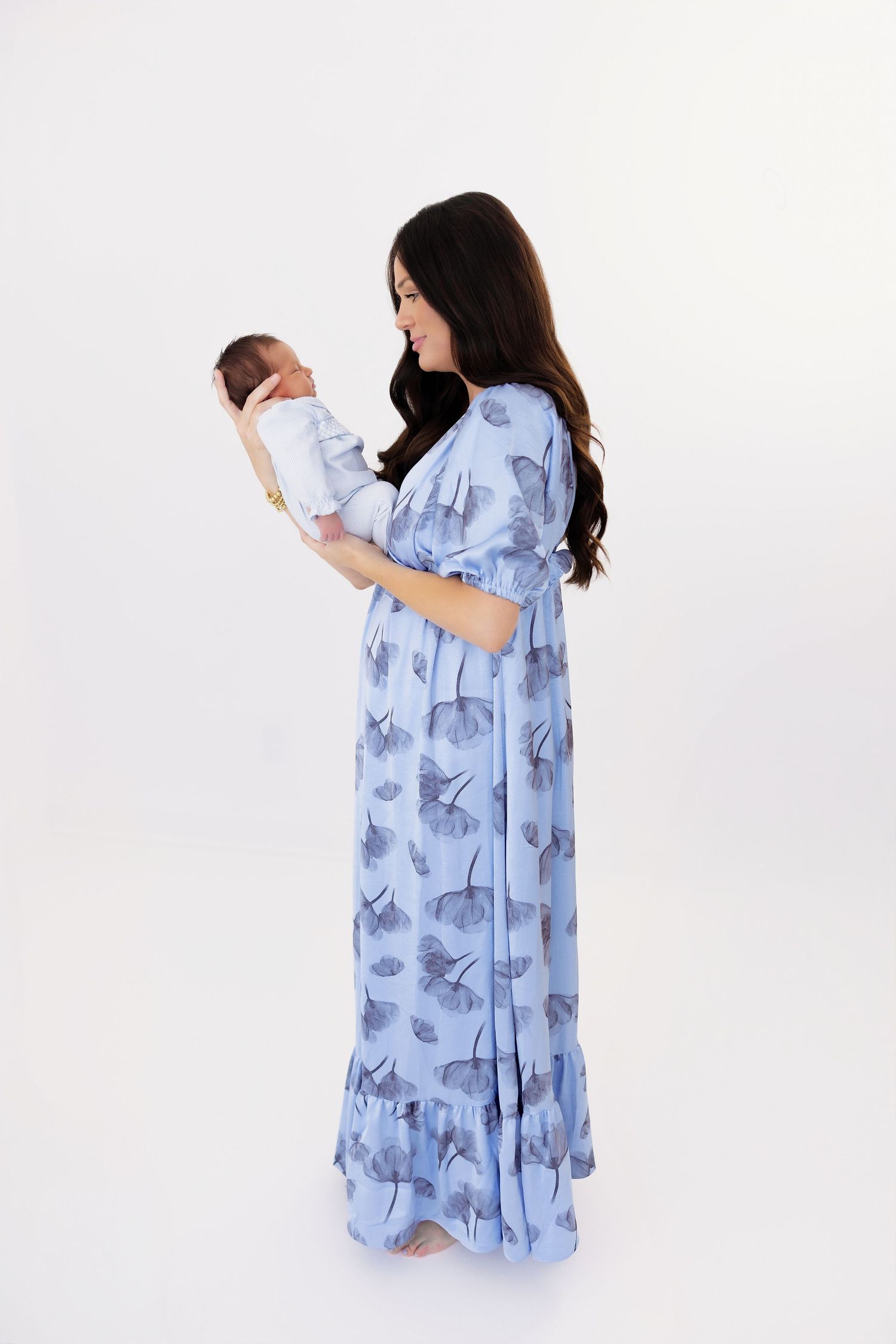 A woman in a blue floral dress holds a newborn baby while standing against a white background.