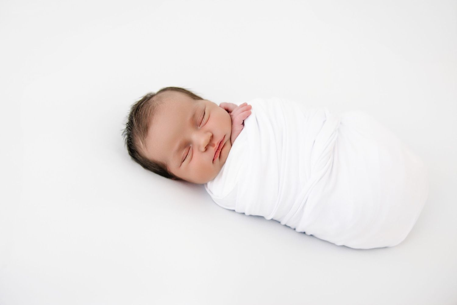 A sleeping newborn baby swaddled in a white blanket lying against a white backdrop.