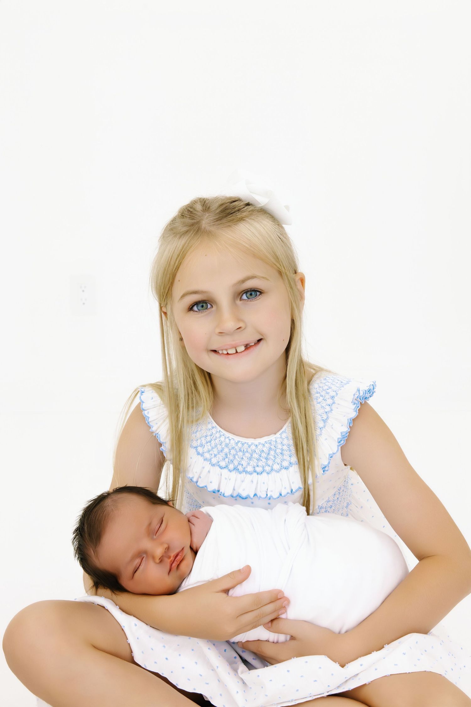 A sweet series showing a young girl in a white dress holding and looking at a sleeping newborn.