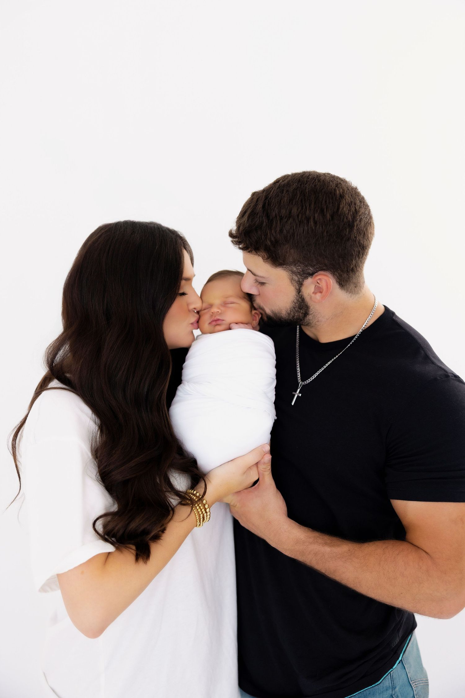 Parents sharing an intimate moment kissing their swaddled baby against a white background.