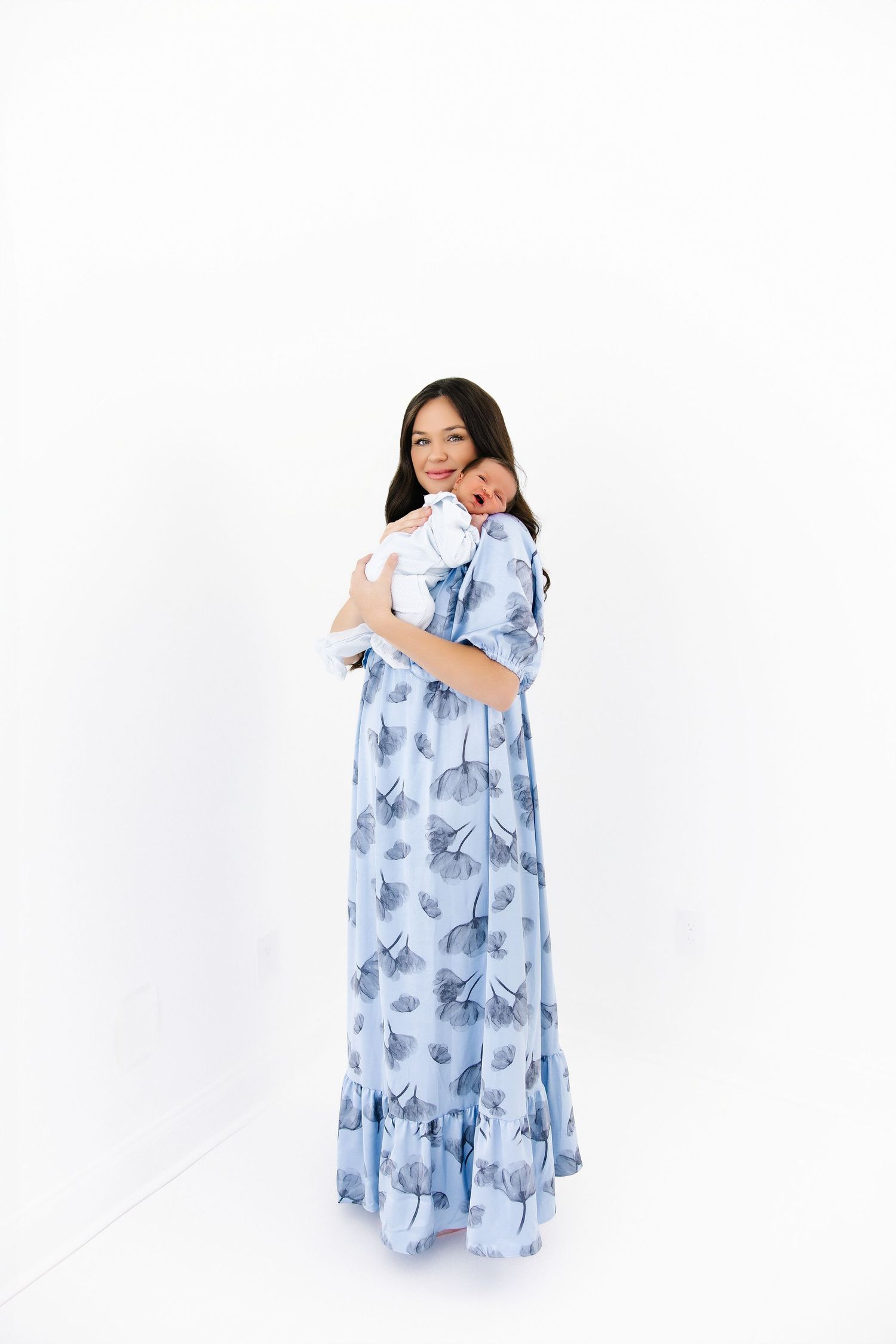 A woman wearing a flowing blue floral dress poses against a white backdrop in a portrait series.