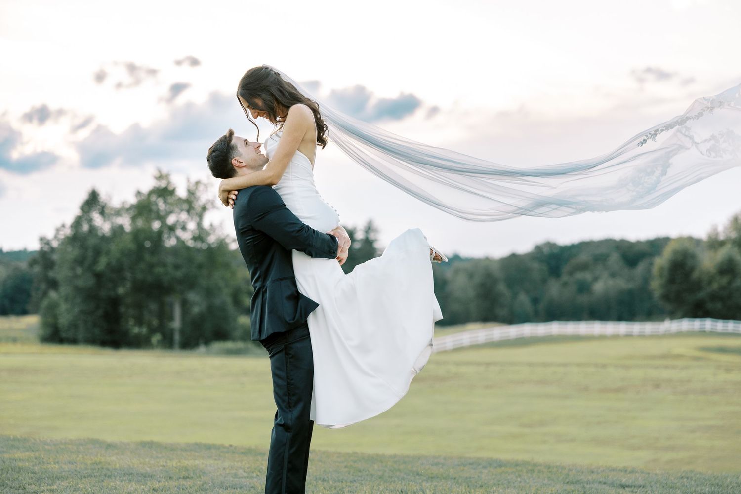 A romantic outdoor wedding photo of a couple in a grassy field with flowing veil during sunset.