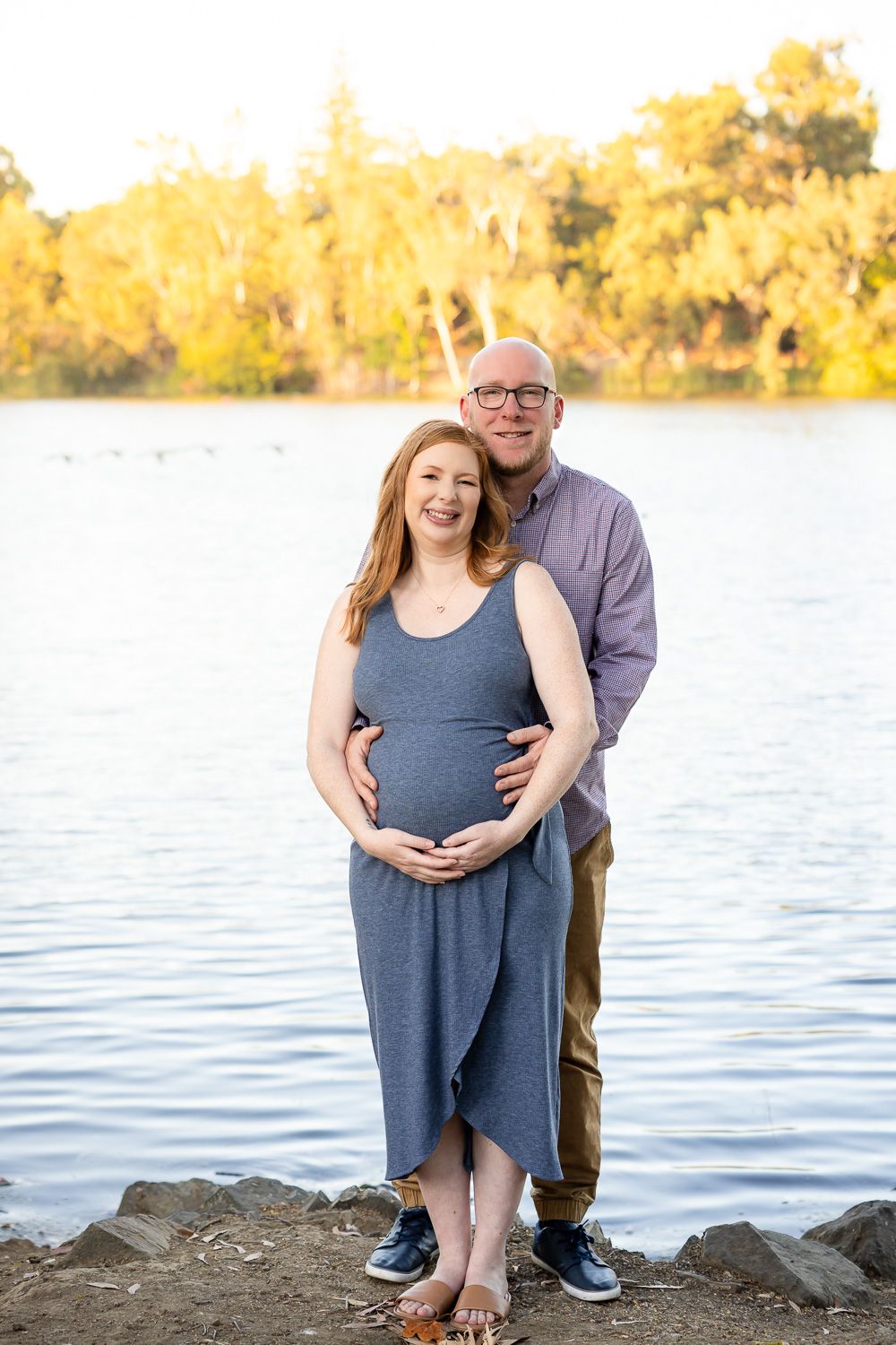 A couple poses together by a lake during autumn with golden trees in the background for their petite maternity photoshoot