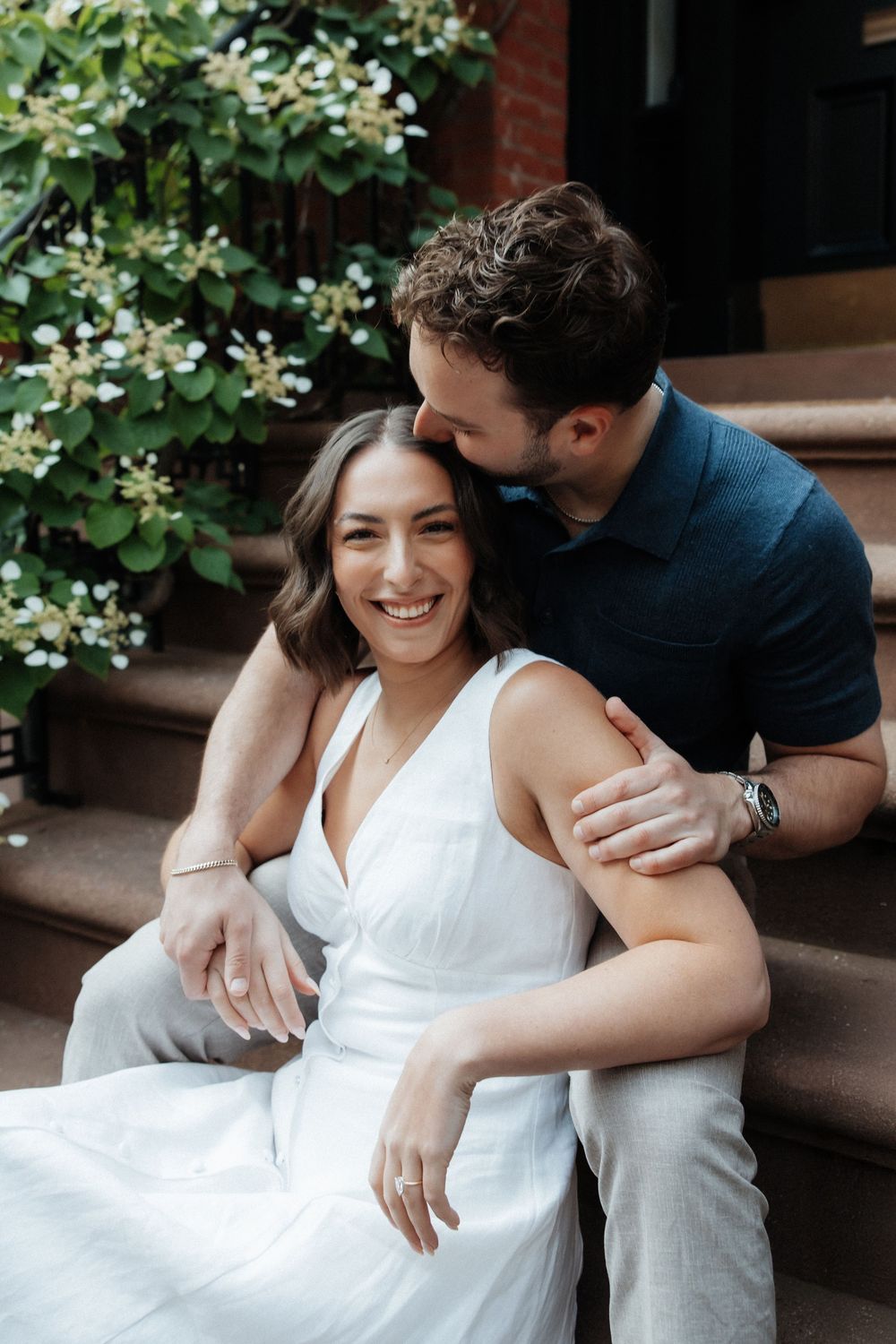 A couple embraces on brownstone steps surrounded by green foliage in an urban setting.
