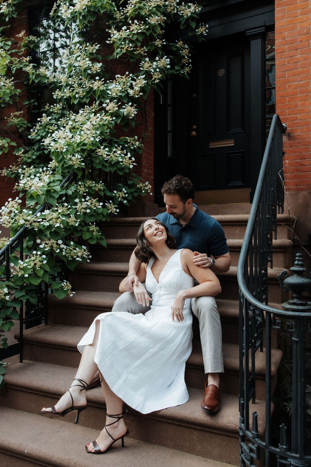 A couple in white and black outfits sits intimately on brownstone steps surrounded by greenery.