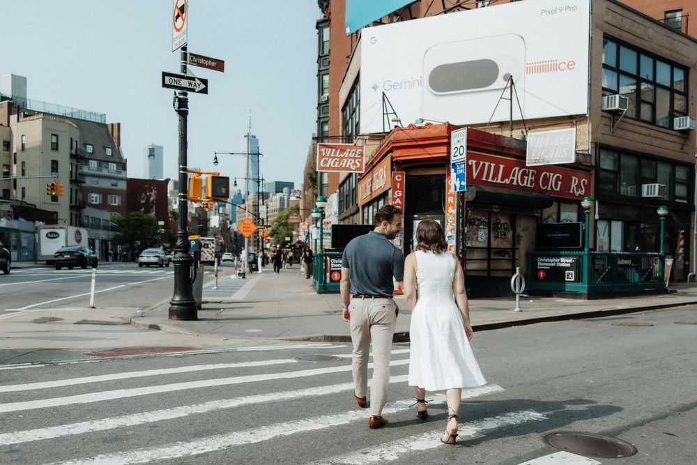 People walk across a city intersection as shops and buildings line a busy urban street in summer weather.