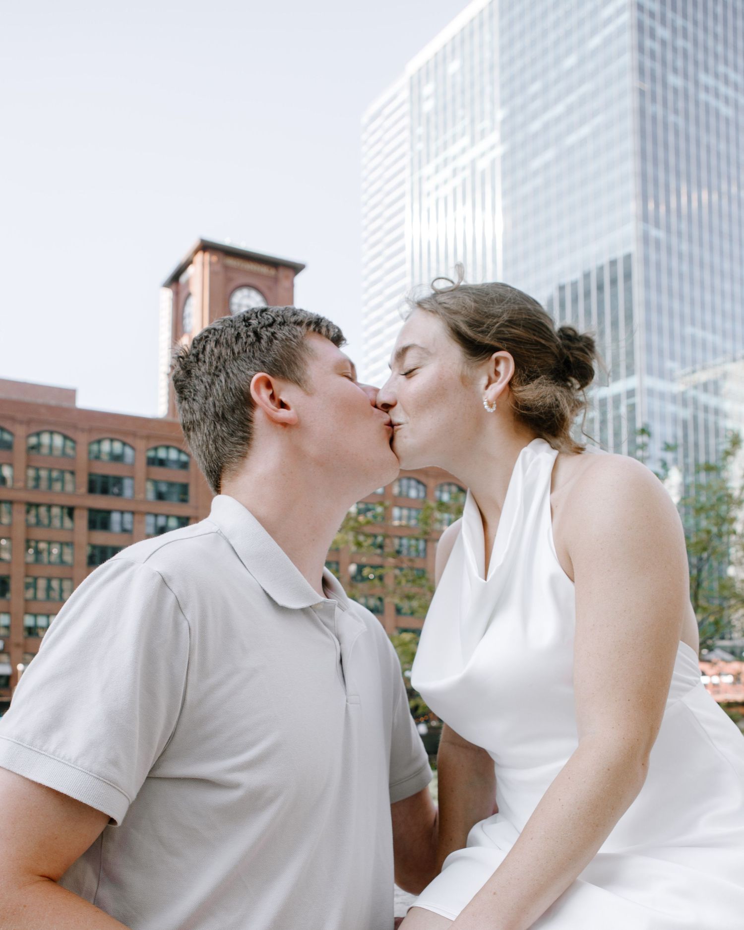 A couple kisses while standing in front of skyscrapers and brick buildings in an urban setting.