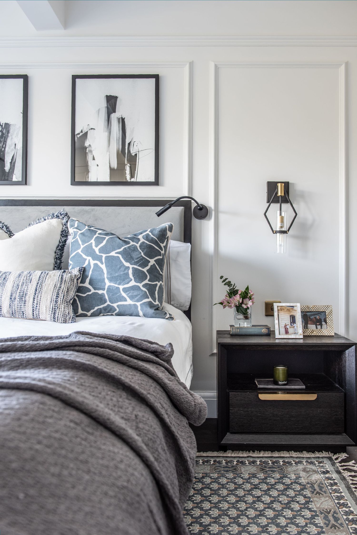 Modern bedroom with grey bedding, blue patterned pillows, black and white wall art, and geometric area rug. Andrew Martin cushions