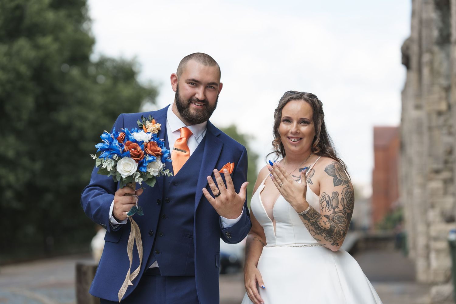 A couple in wedding attire smiling and walking together outdoors, with the groom holding a blue and white bouquet.