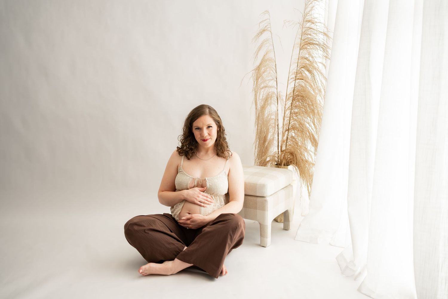 A pregnant woman sits cross-legged on a white floor wearing brown pants and white tank top, near pampas grass decor.