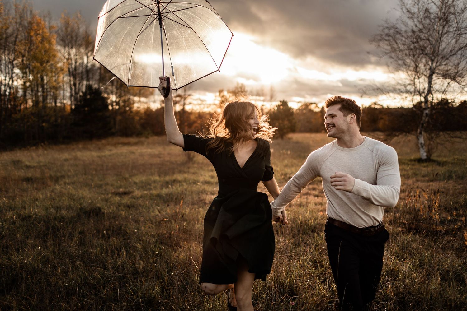 Couple frolicking in a field at sunset with a white umbrella creating a dreamy atmosphere.