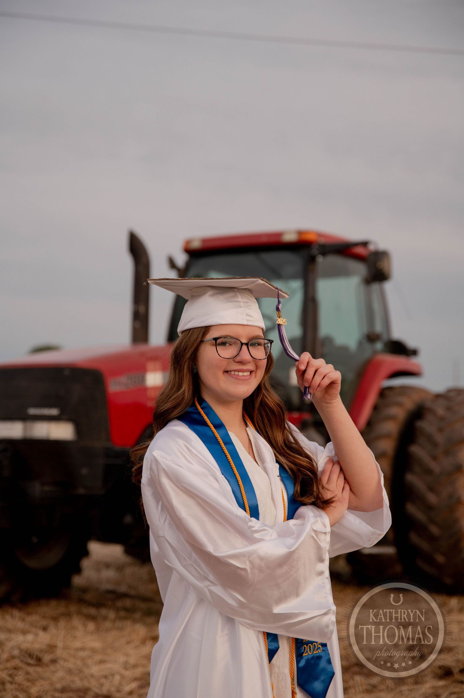 A graduate in white cap and gown stands near a red tractor in a rural field setting during a photo session.