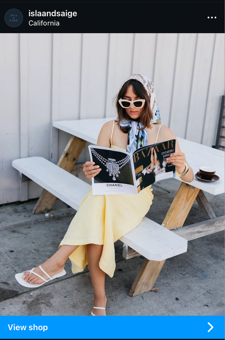 Person wearing sunglasses and a black top relaxes at an outdoor cafe with magazine and coffee in sunny California.