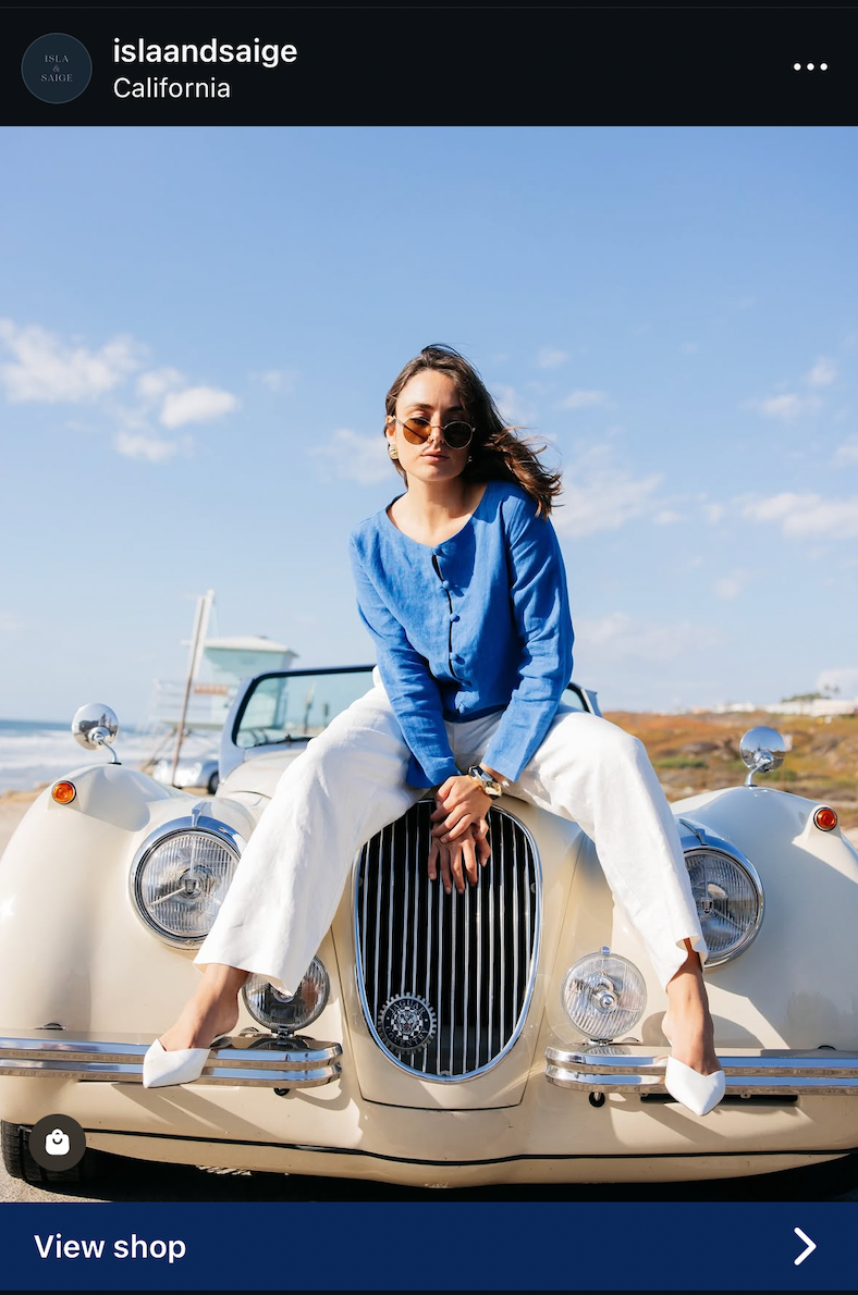 A person in a blue sweater and white pants sits on the hood of a vintage cream-colored Jaguar car at the beach.