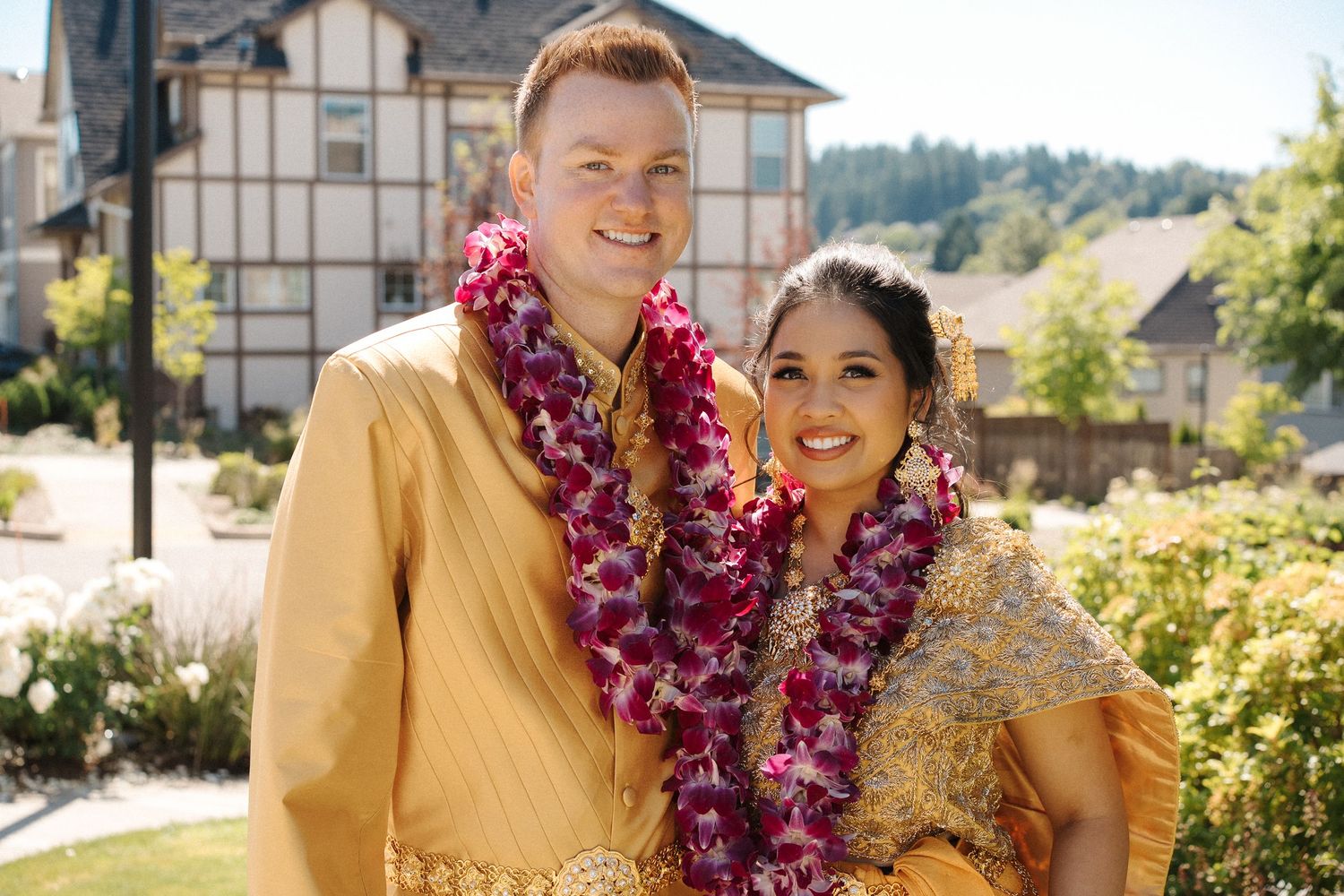 Two people wearing vibrant purple and pink Hawaiian leis smile outdoors on a sunny day with buildings in the background.