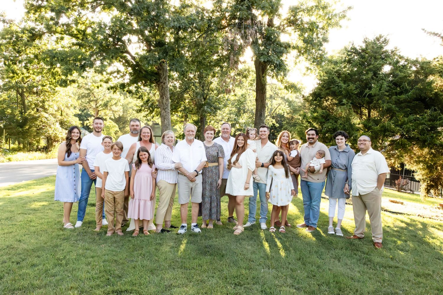 Large extended family group wearing pastel and neutral colored outfits poses together on a sunny lawn for formal portraits.
