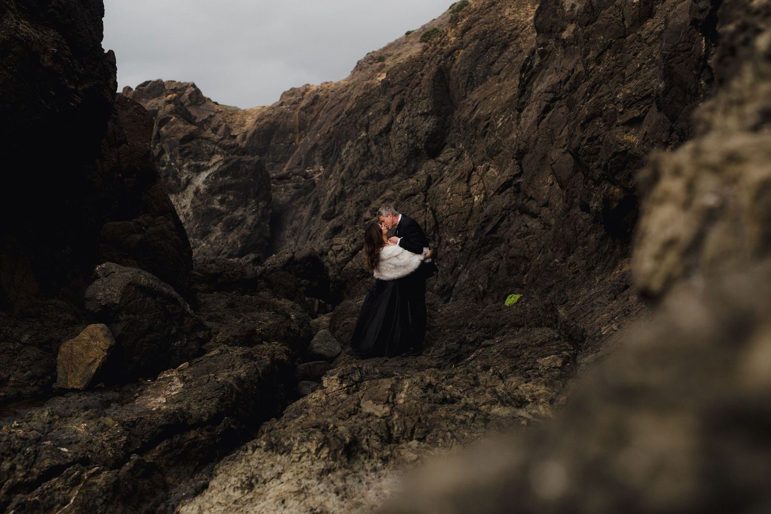 A couple in dark formal attire stands on cliff edge overlooking dramatic rocky coastline and misty ocean view.