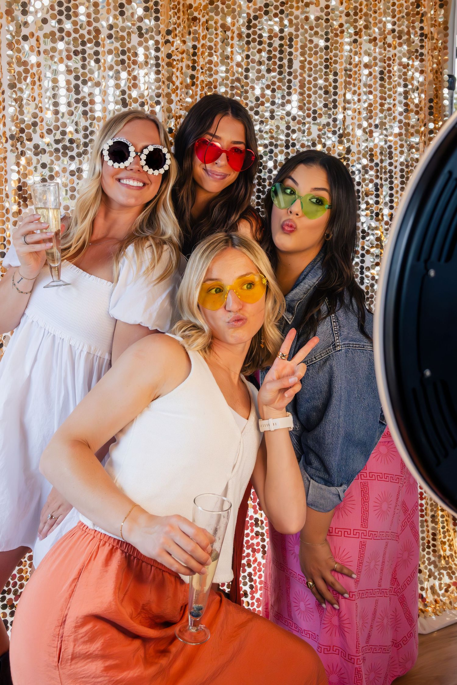 Group wearing colorful sunglasses pose together against a sparkly gold sequin backdrop at a party celebration.