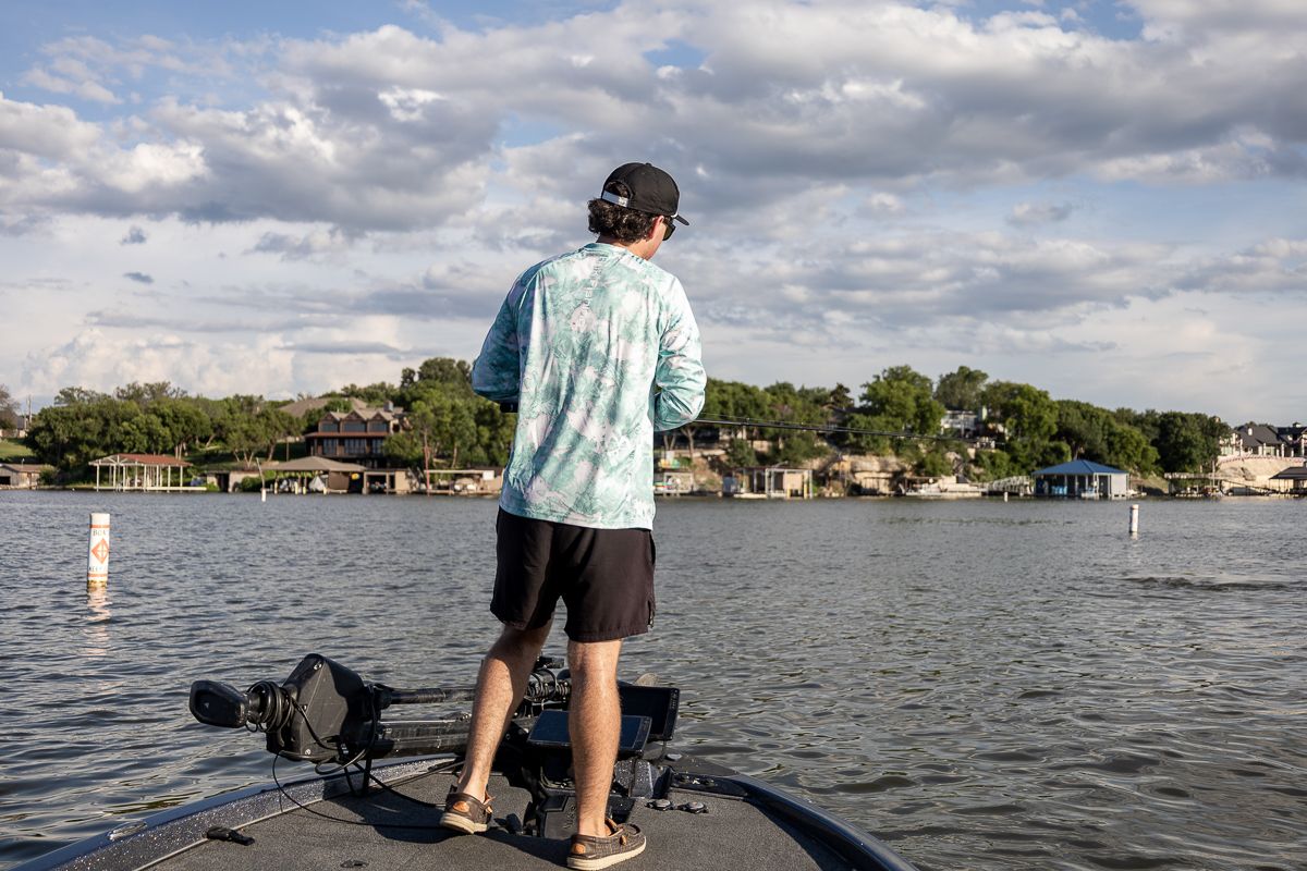 Angler stands on a boat deck overlooking a scenic lake with waterfront homes and cloudy skies in the background.