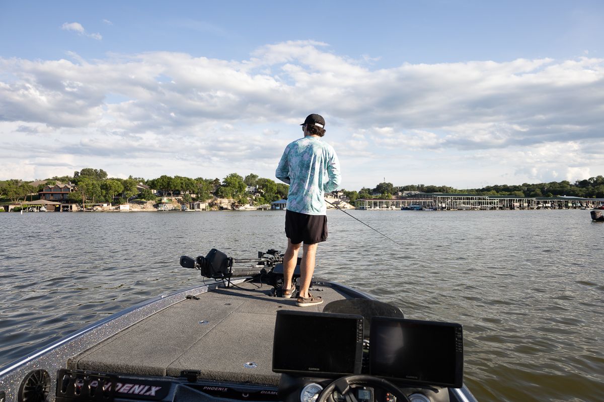 Fisher stands on the bow of a bass boat on calm waters with lakeside houses and docks visible.