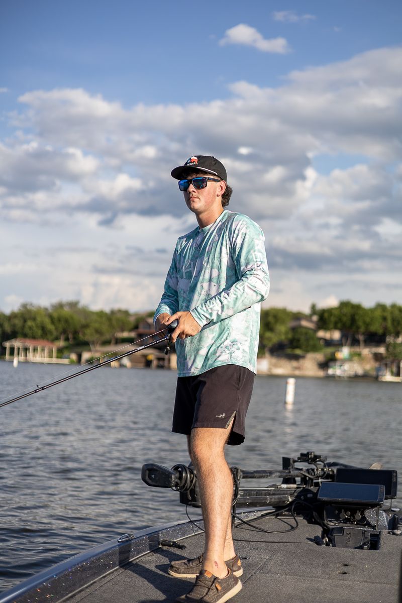 Angler in light blue shirt and shorts fishing from the deck of a bass boat on calm waters.