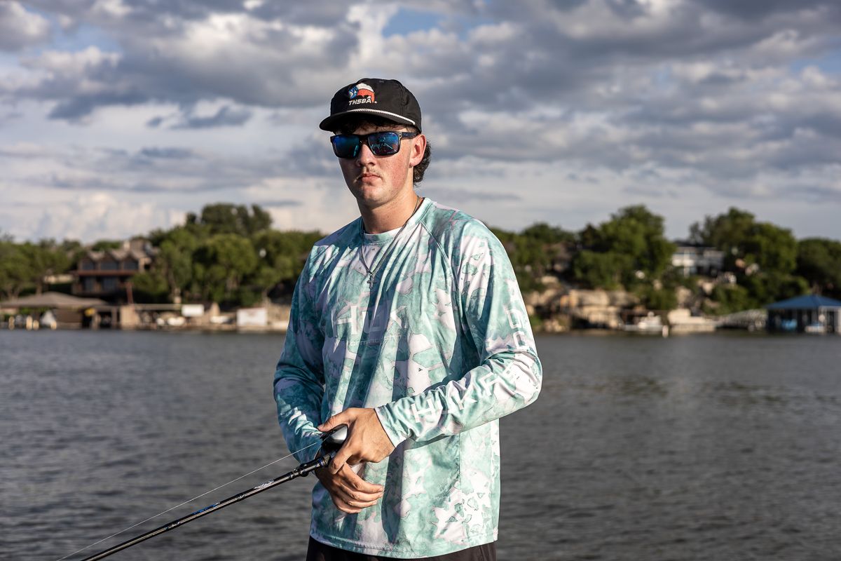 Angler wearing protective sun gear stands ready with fishing rod against scenic waterfront backdrop with cloudy skies.