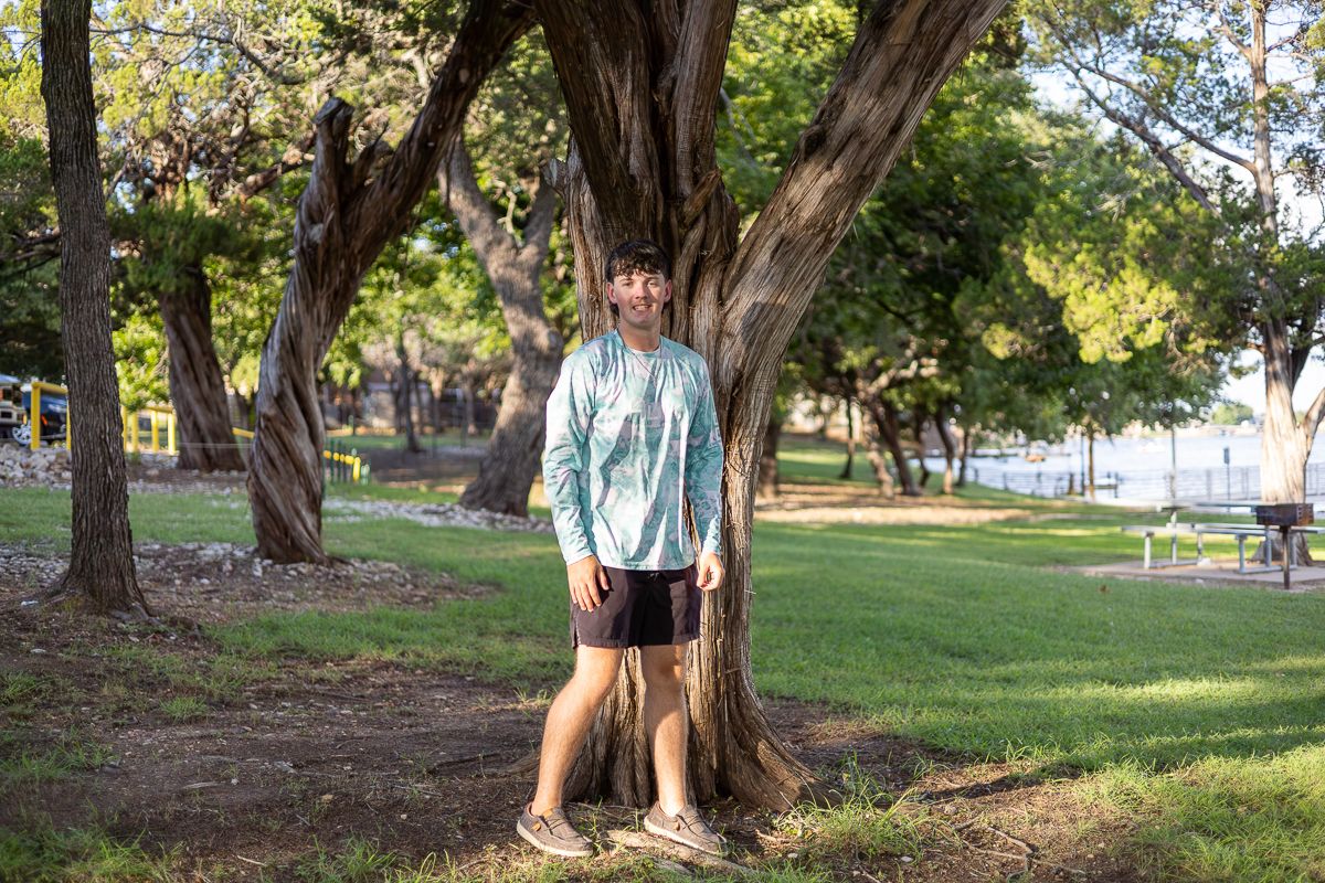 Person in light colored clothing standing next to large tree trunk in grassy waterfront park.