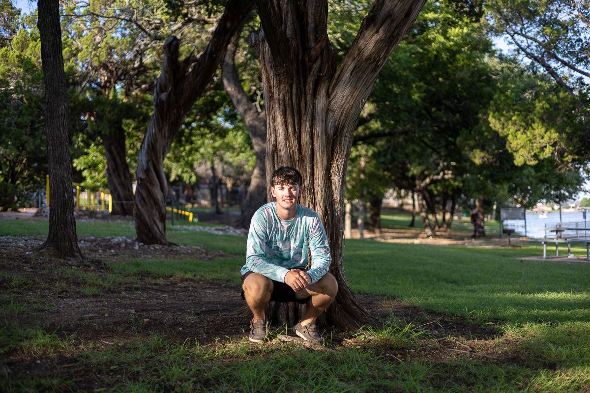 Outdoor lakeside scene with person sitting under large shady trees in a park setting.