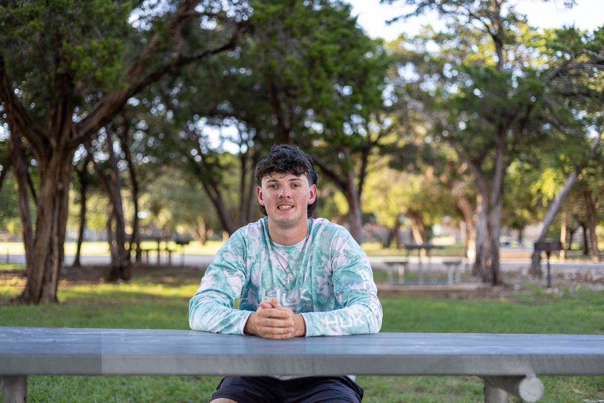 Person wearing light blue sweater sits at outdoor table in a sunny park with trees in background.