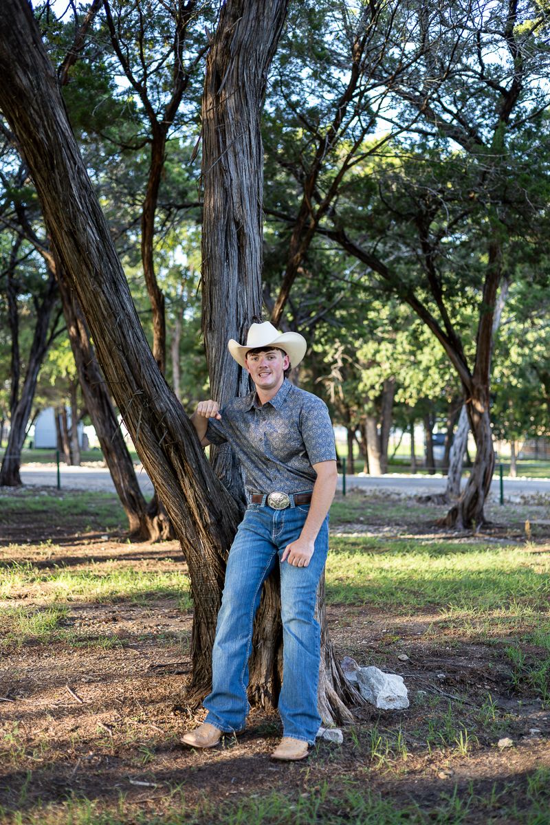 Country western style with cowboy hat and jeans standing casually against a tree trunk in a park setting.