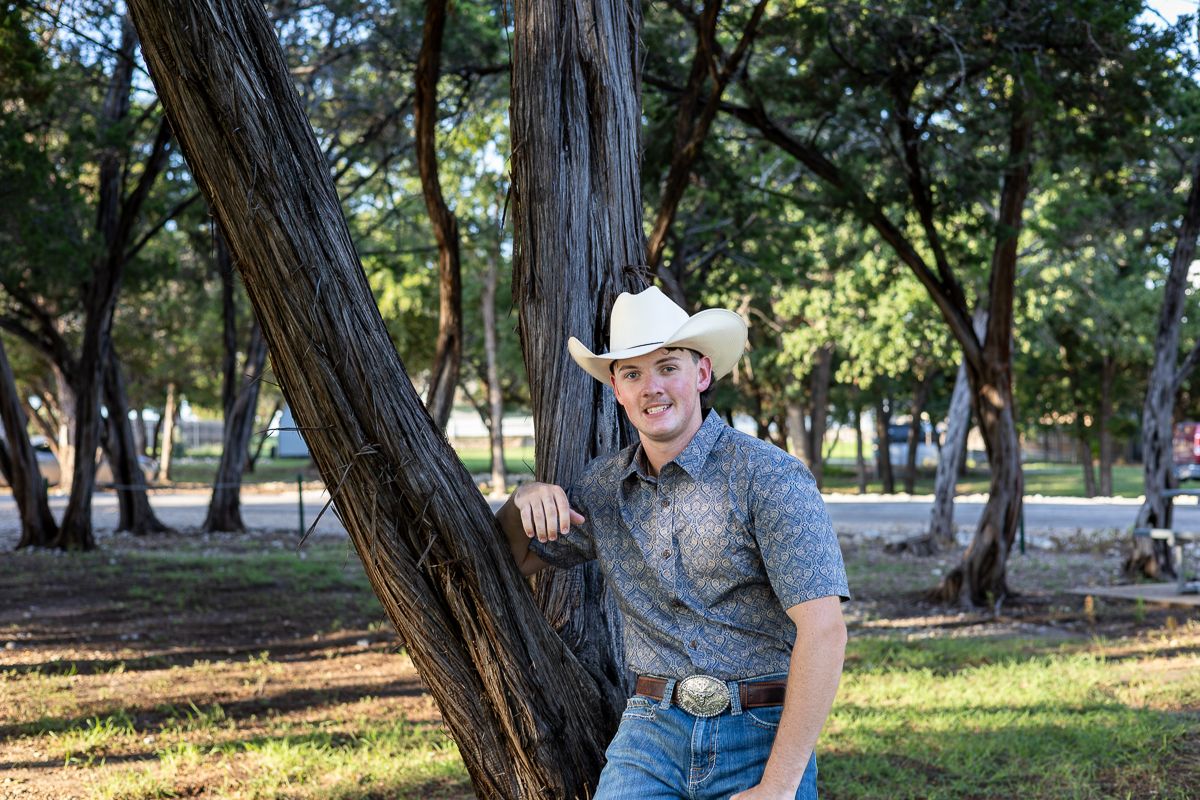 Person in western attire leaning casually against a tree trunk in a sunlit park setting.
