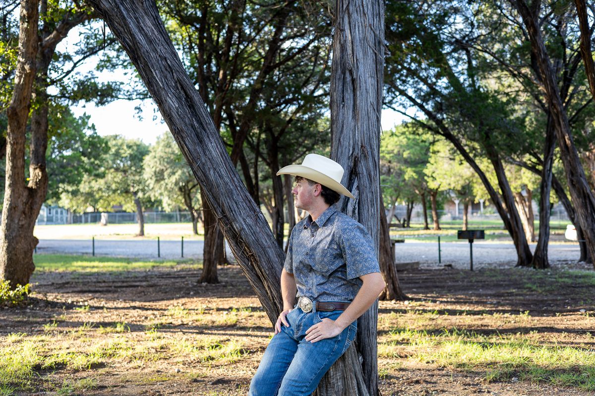 A person in denim and cowboy boots leans against a tree trunk in a sunlit wooded area.