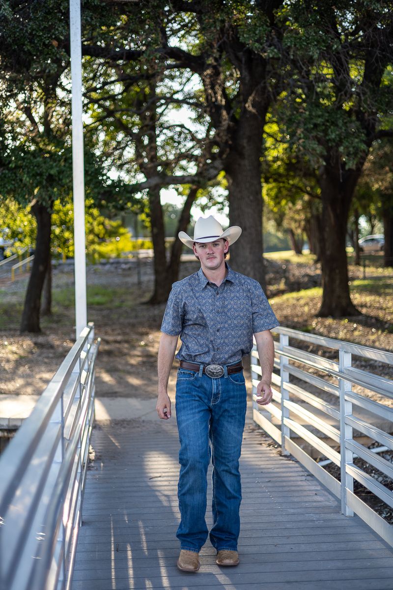 A figure in a cowboy hat and western attire walks along a wooden boardwalk surrounded by trees in warm sunlight.
