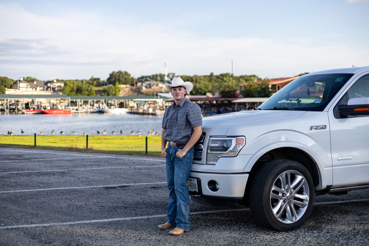 White pickup truck parked in gravel lot against scenic waterfront and residential background.