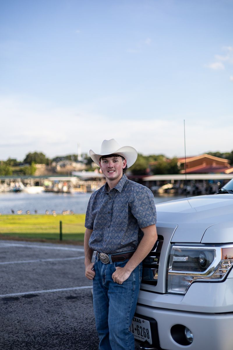 A person in a cowboy hat stands next to a white pickup truck by the waterfront during daytime.
