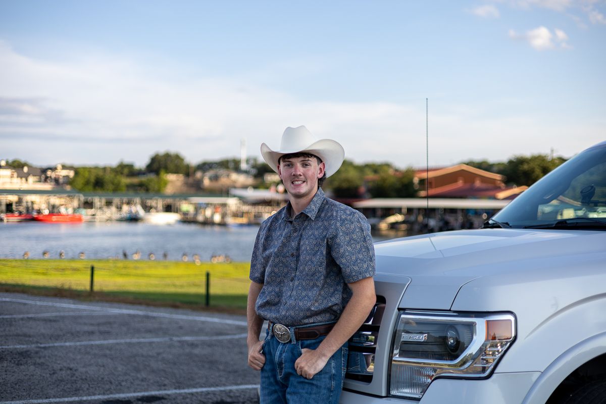 A person in a cowboy hat and gray shirt stands casually next to a silver pickup truck parked by a waterfront.