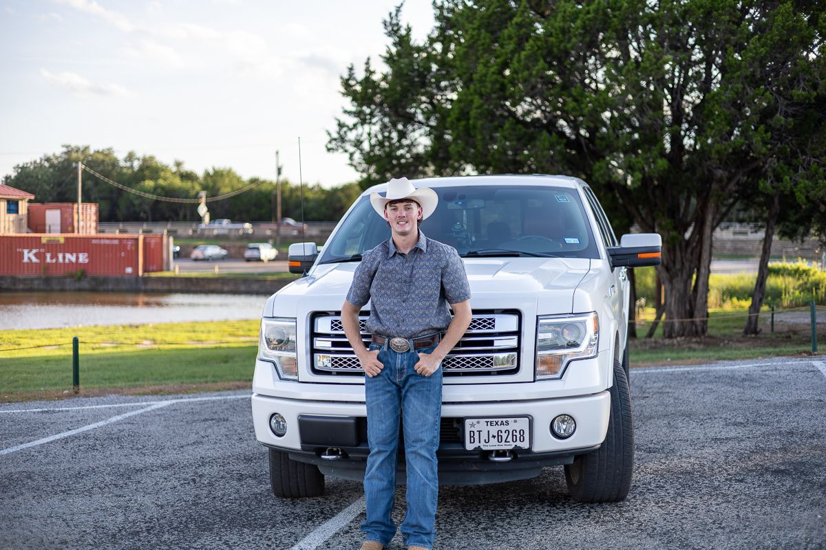 A person in western attire poses in front of a white Ford F-150 pickup truck in a parking lot.