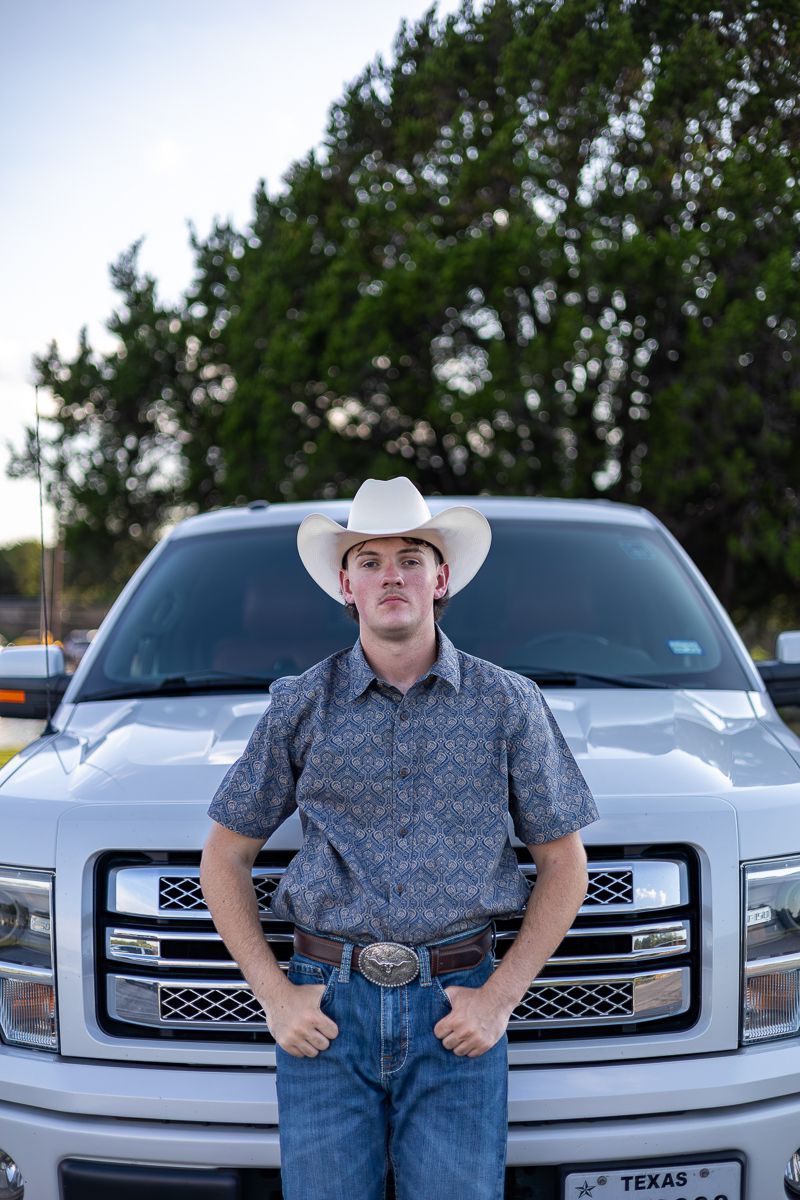 Person in cowboy hat and western attire stands in front of silver pickup truck with trees in background.