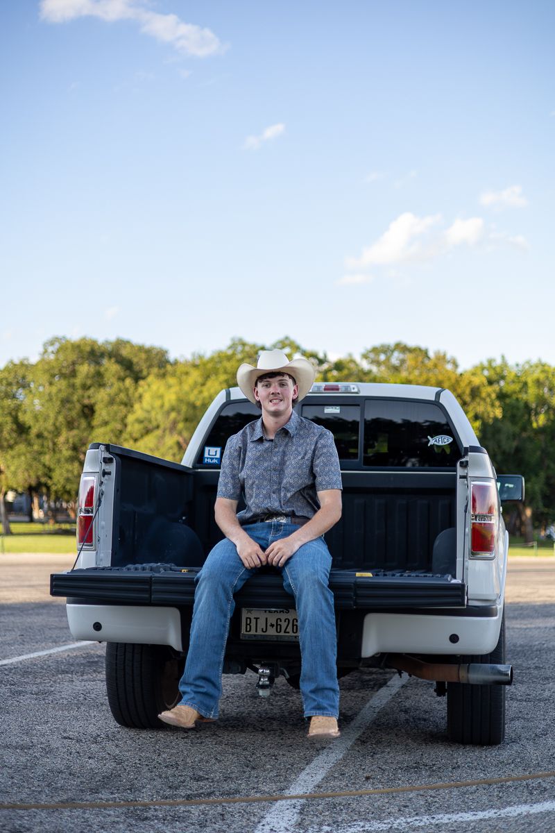 A relaxed figure in western wear sits on the tailgate of a white pickup truck in a sunny parking lot.
