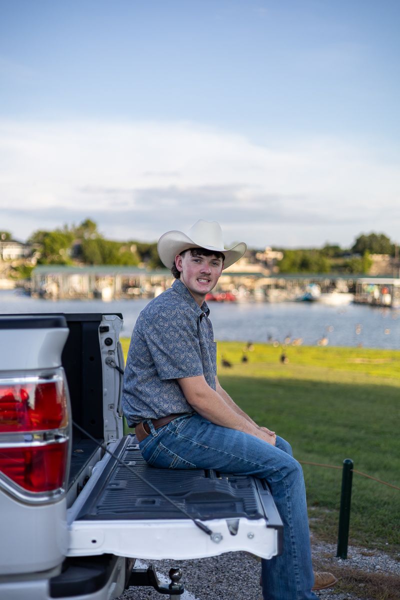 A person in a cowboy hat sits casually on the tailgate of a pickup truck with a marina and boats visible in the background.