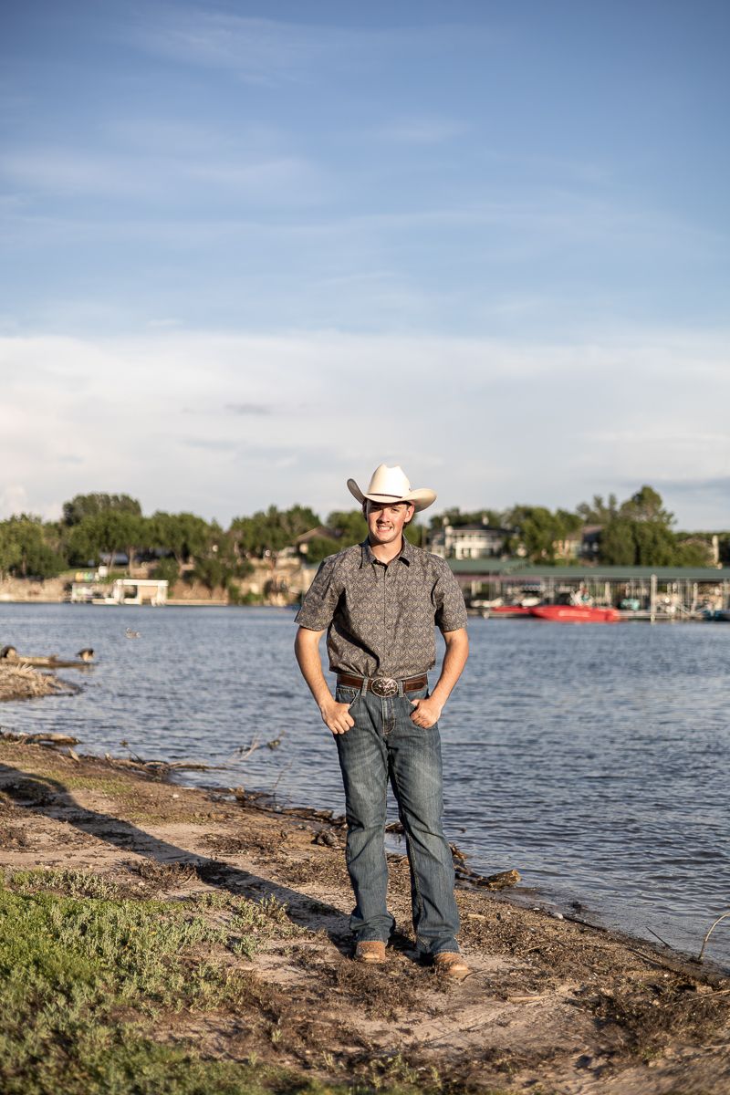 Person in western wear stands on rocky shoreline with boats and lake in the background.