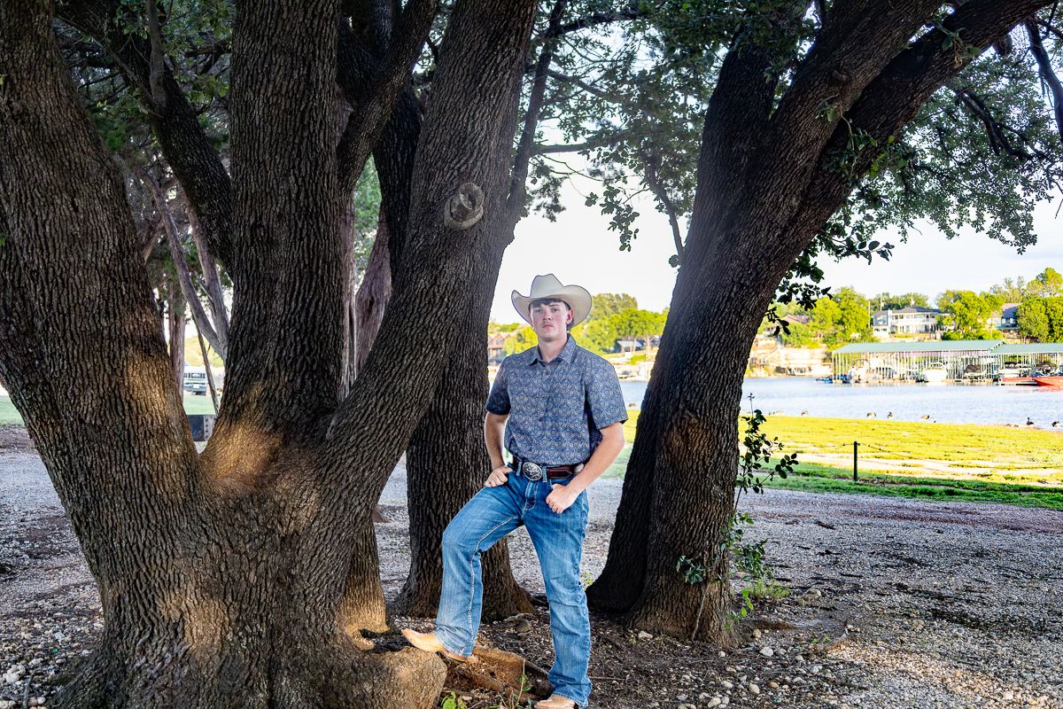 A person stands casually against large tree trunks in a sunny outdoor park setting.