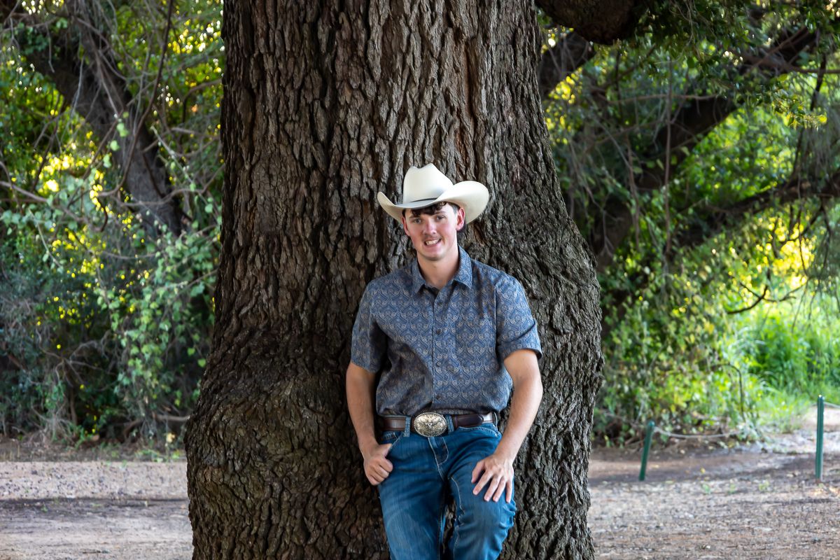 Person in cowboy hat poses against large tree trunk with dappled sunlight filtering through branches.