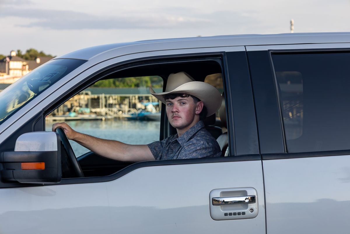 A driver wearing a cowboy hat sits behind the wheel of a silver pickup truck with a marina visible in the background.