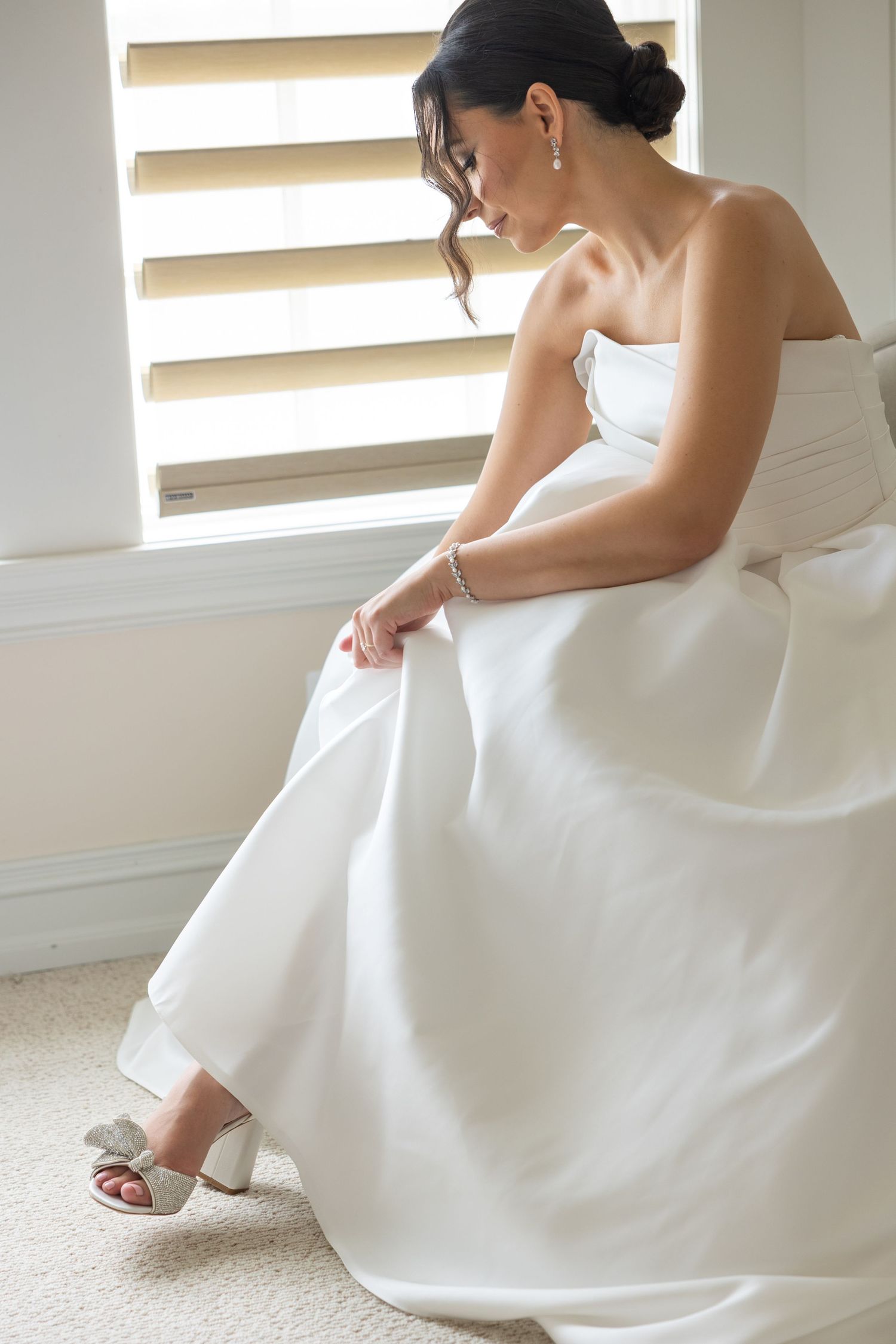 Bride in elegant white wedding gown sits by window while adjusting her silver high heel shoe.