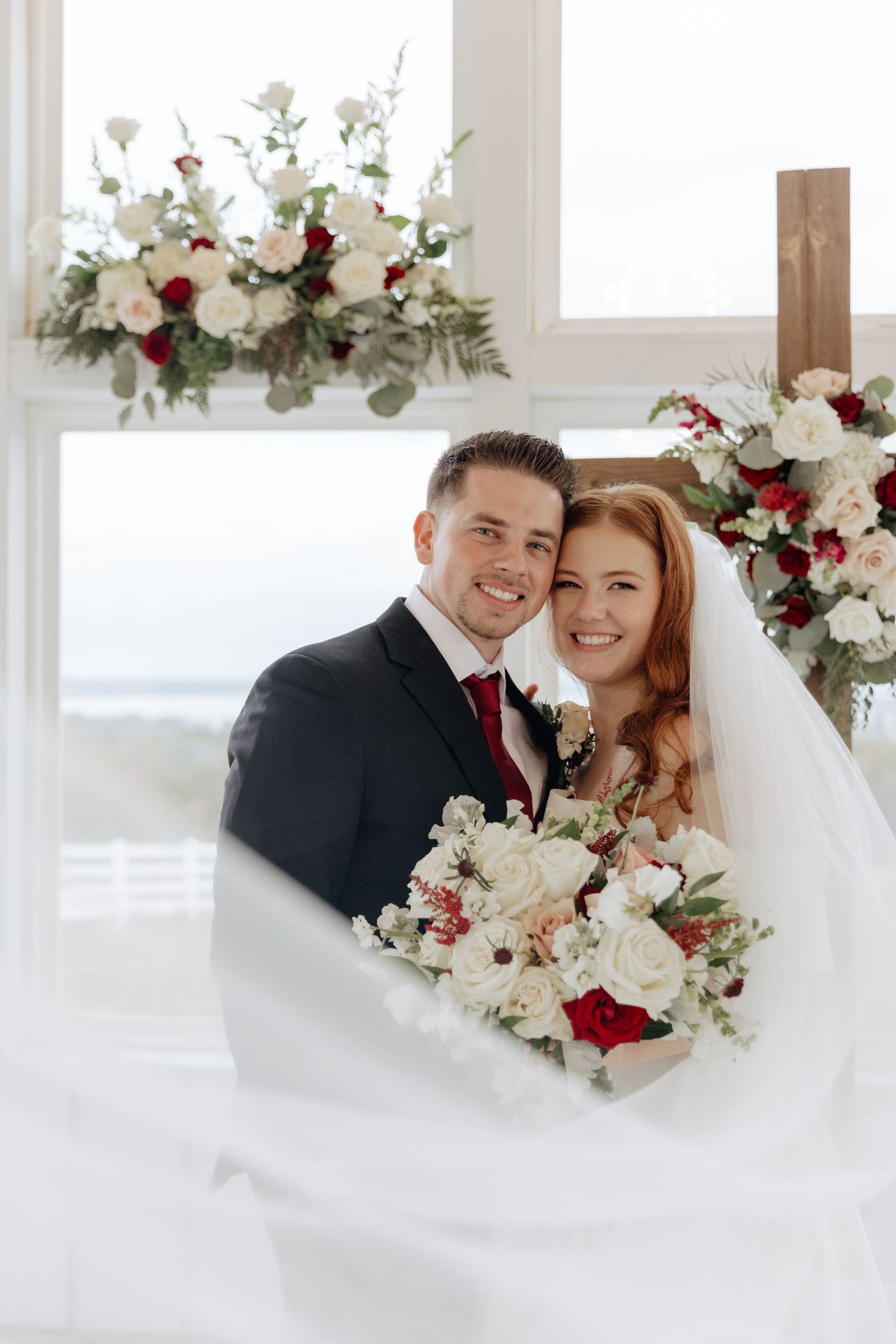 Romantic wedding couple embraces near floral arrangements featuring red and white roses in bright window-lit space.
