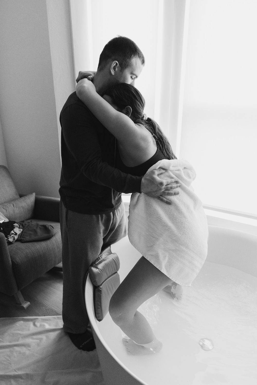 Black and white photo captures intimate moment during labor as partner supports woman in labor leaning over birthing tub.