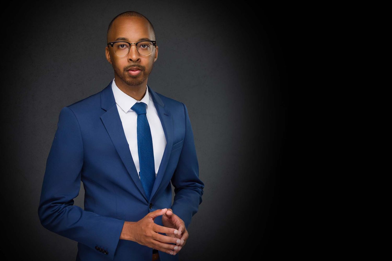 Professional portrait of someone in a sharp navy blue suit with white shirt, blue tie and glasses against dark background.